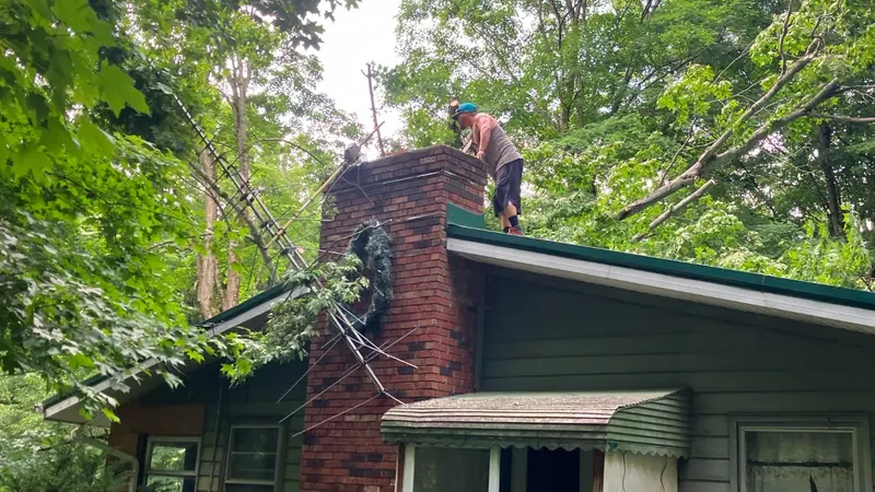 A crew member on a roof cutting storm-fallen limbs away beside a brick chimney