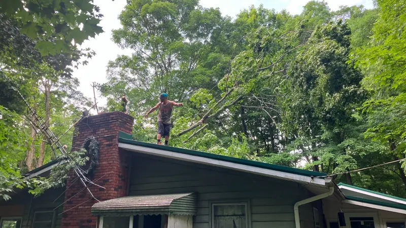 A crew member on a house roof clearing a storm-fallen tree