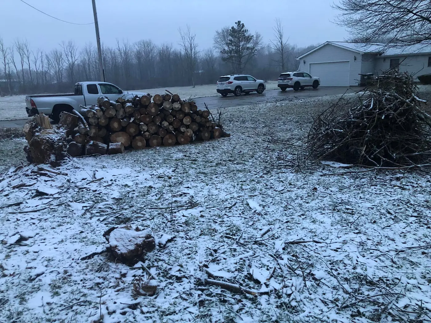 A cleared property under snow with cut logs stacked, brush piled to one side, and a stump cut flush to the ground